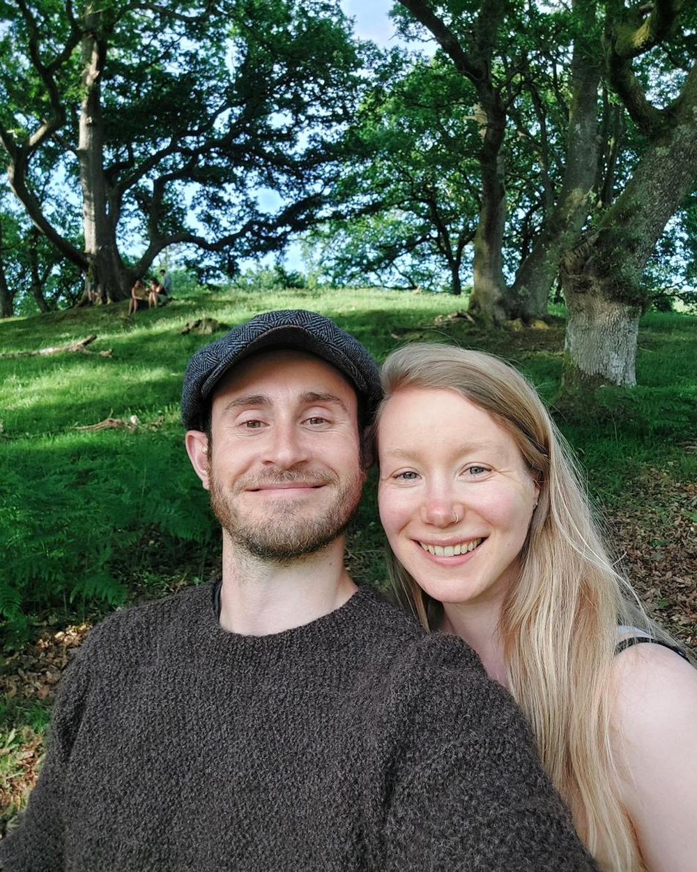 Jamie and Bex smiling together beneath oak trees on a green hillside.